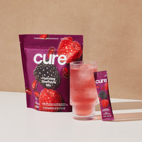Cure Hydration Electrolyte Mix package with images of berries and pomegranate, next to a glass of pink beverage and a single-serve stick pack labeled 'cure'