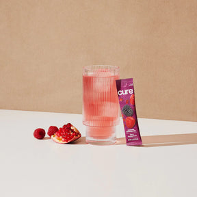 Glass of pink drink with ice, Cure Berry Watermelon supplement stick, raspberries, and pomegranate seeds on a white surface with beige background