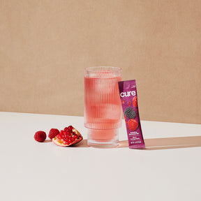 Two ribbed glasses filled with pink liquid, a pomegranate half, scattered raspberries, and a purple packet labeled 'cure Berry Pomegranate' on a white surface with a beige background.