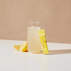 Glass of lemonade with ice, lemon wedge, and yellow 'cure' packet labeled 'Berry Pomegranate/Lemonade Bundle'