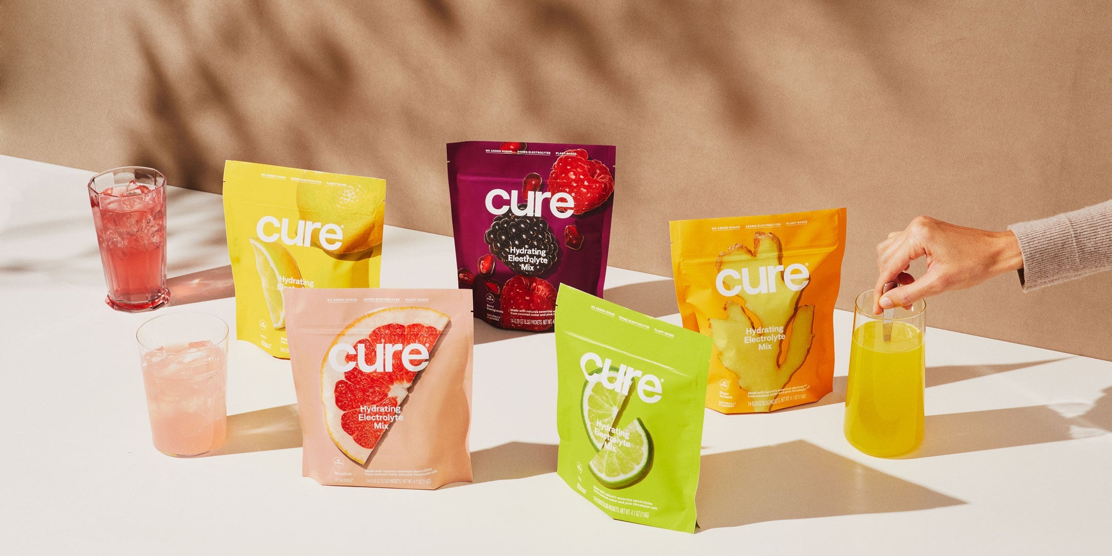 Five colorful packets of Cure Hydrating Electrolyte Mix in lemon, raspberry, grapefruit, lime, and ginger flavors, with glasses of mixed drinks and a hand stirring the yellow drink.