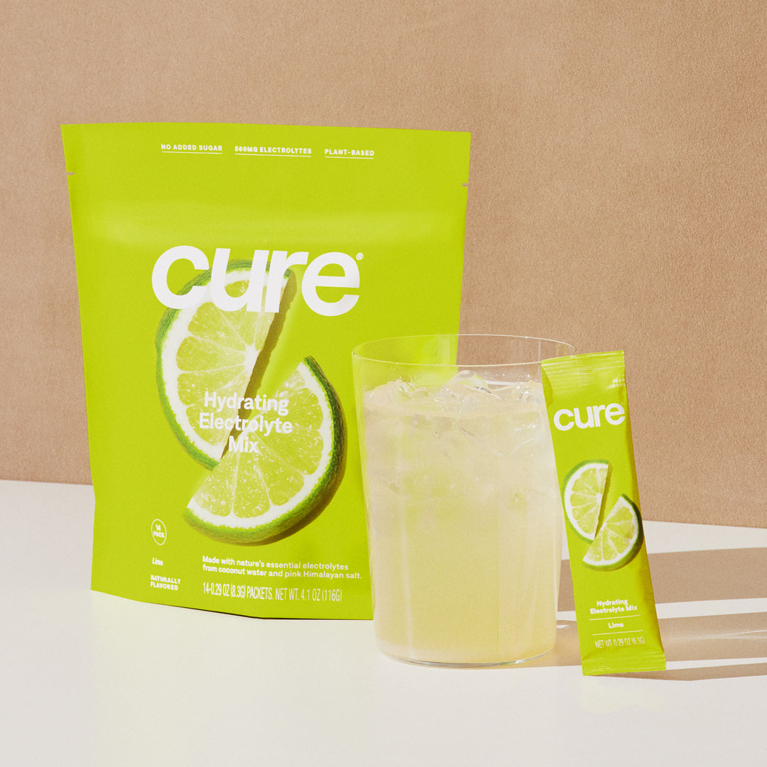 Package of Cure Hydrating Electrolyte Mix in Lemon Lime flavor, with a glass of the prepared drink and a single stick pack beside it