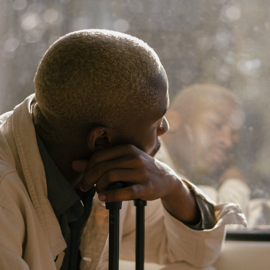 A person with short light brown hair resting their chin on their hand, looking out a window with their reflection visible.