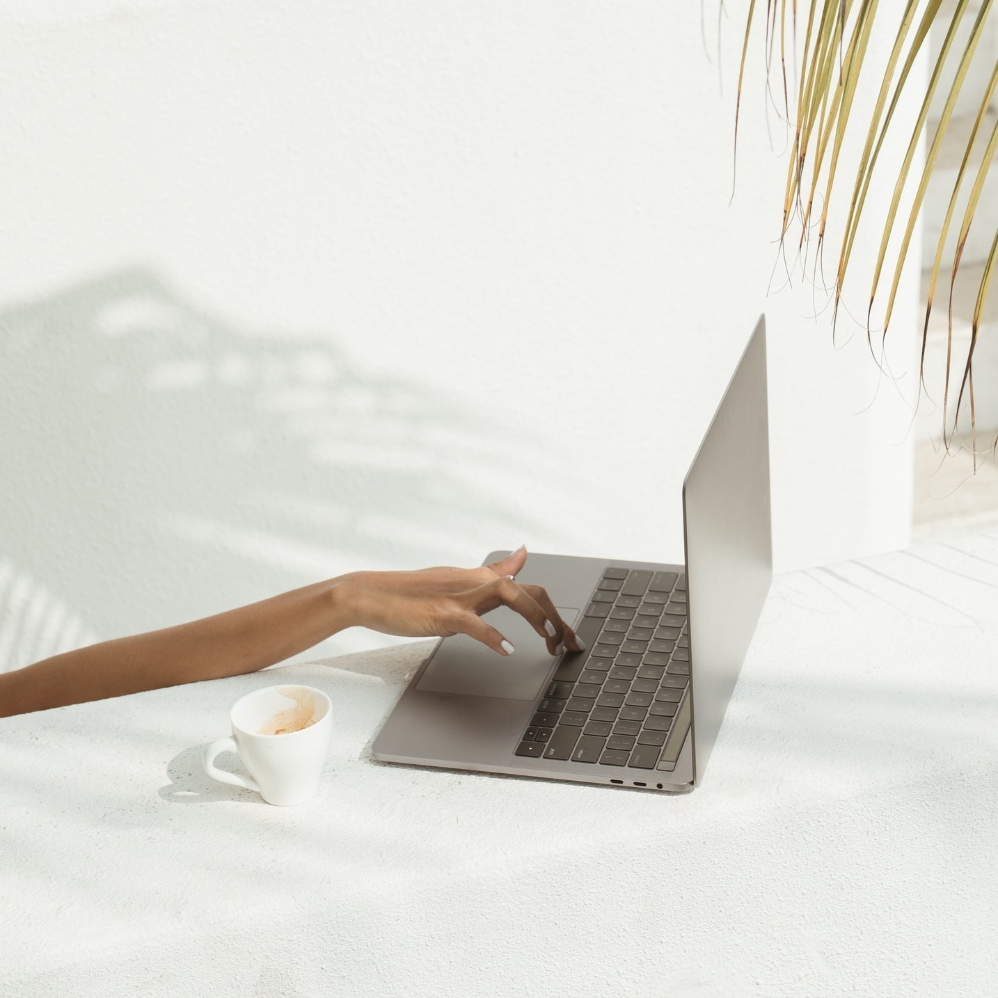 A hand typing on a laptop keyboard next to a white coffee cup on a white surface with shadows from nearby plants.