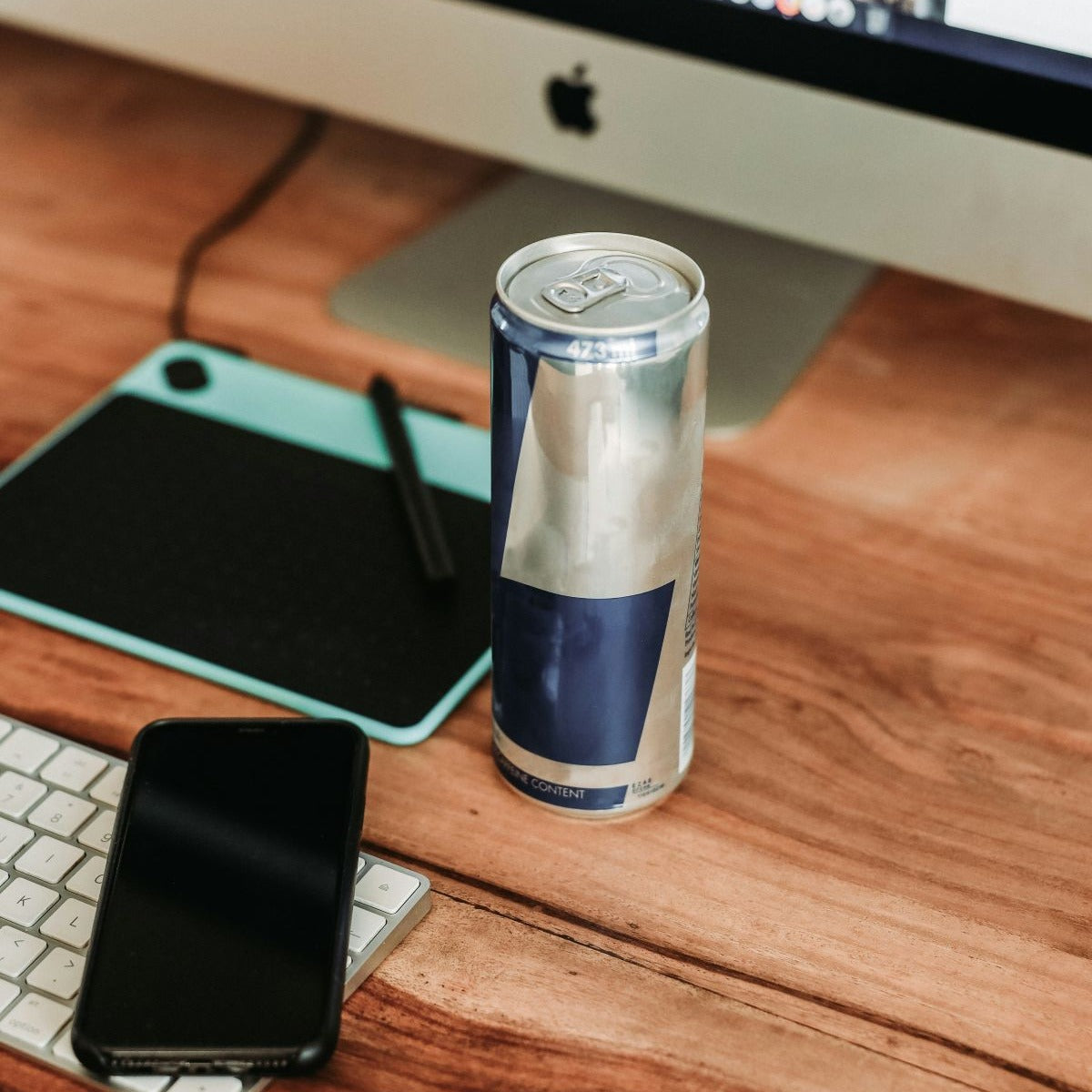 Energy drink can on a wooden desk near a keyboard, smartphone, and tablet with stylus.
