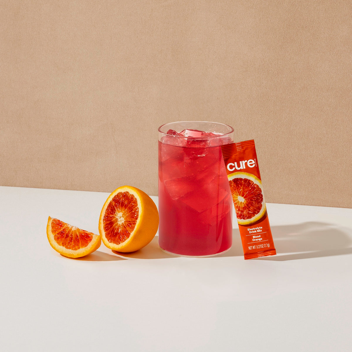 Glass of red blood orange drink with ice, blood orange fruit halves, and a 'cure' drink mix packet
