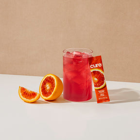 Glass of red blood orange drink with ice, blood orange fruit halves, and a 'cure' drink mix packet