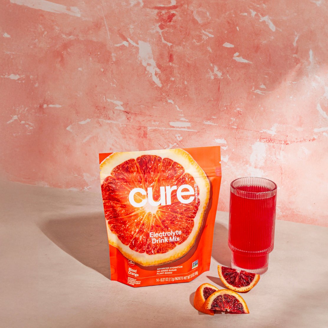 Cure Electrolyte Drink Mix package with blood orange image, a glass filled with red drink, and two blood orange slices on a pink textured background