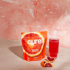 Cure Electrolyte Drink Mix package with blood orange image, a glass filled with red drink, and two blood orange slices on a pink textured background