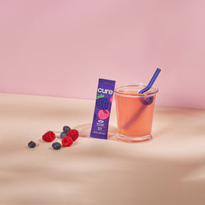 Glass of pink mixed berry drink with a blue straw, a purple box labeled 'cure' with a raspberry image, and scattered blueberries and raspberries on a beige surface.
