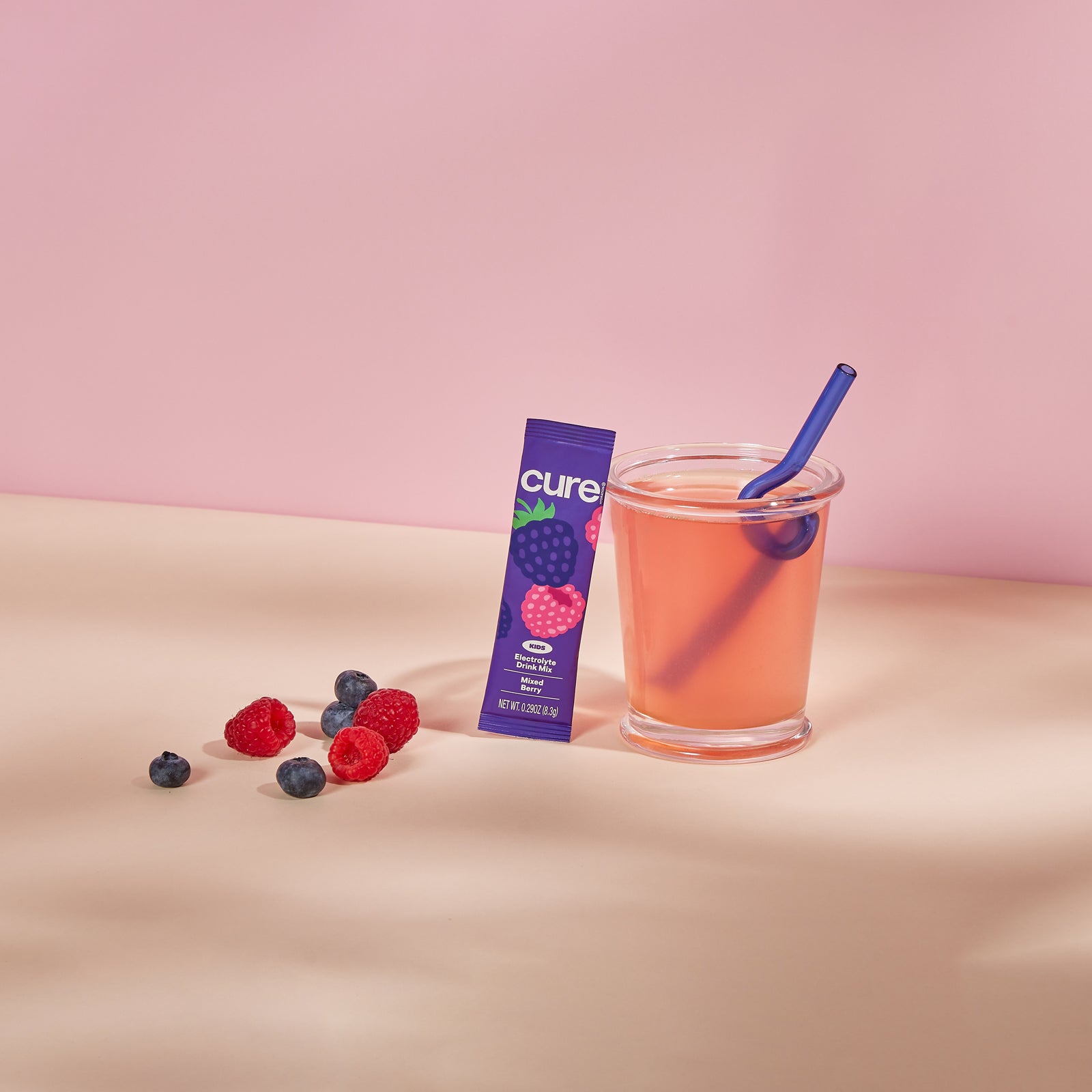 Glass of pink drink with a blue straw next to a purple Cure Kids Variety Pack packet and scattered raspberries and blueberries on a light surface with a pink background