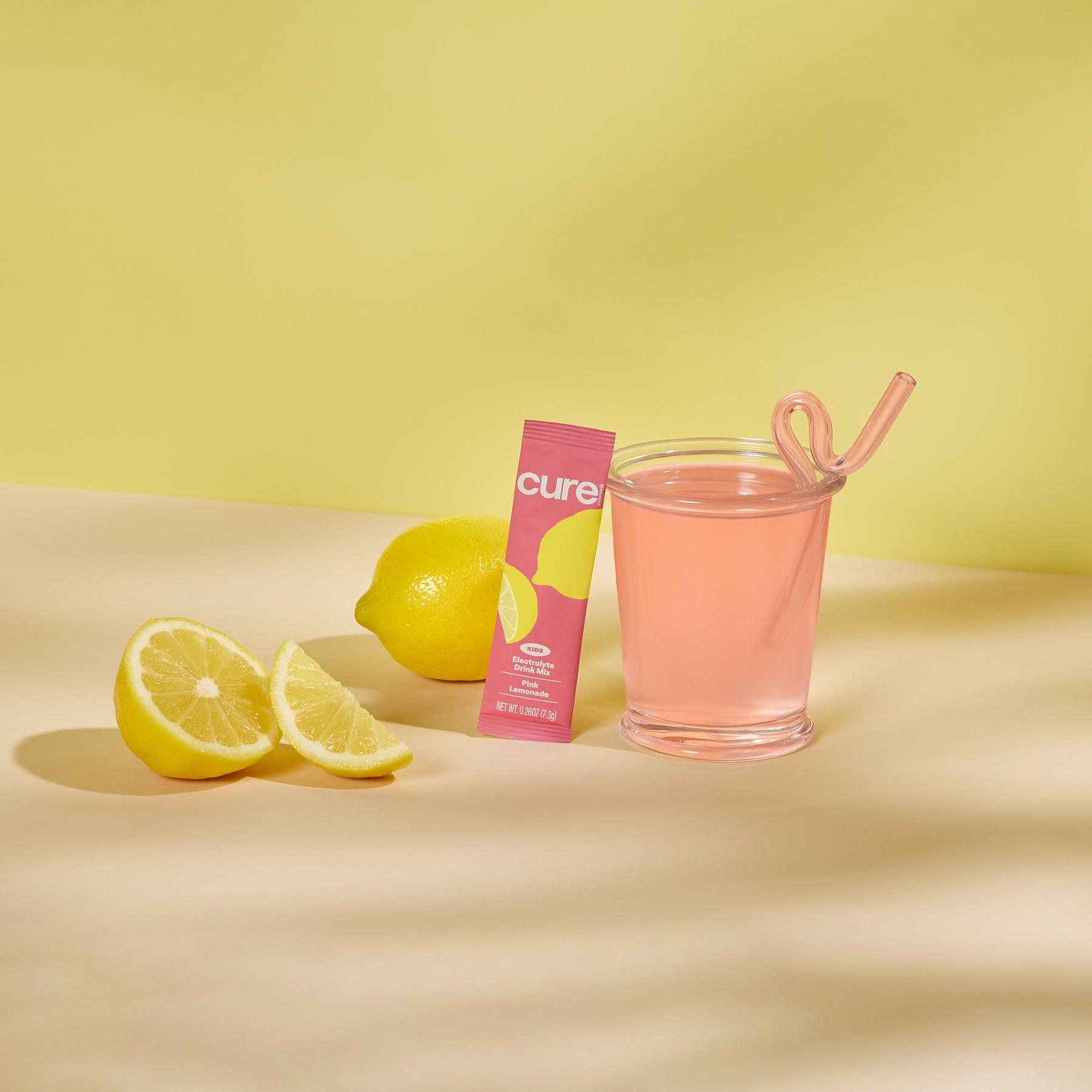 Glass of pink lemonade with a pink straw, a pink Cure product box, and two lemon slices on a yellow background