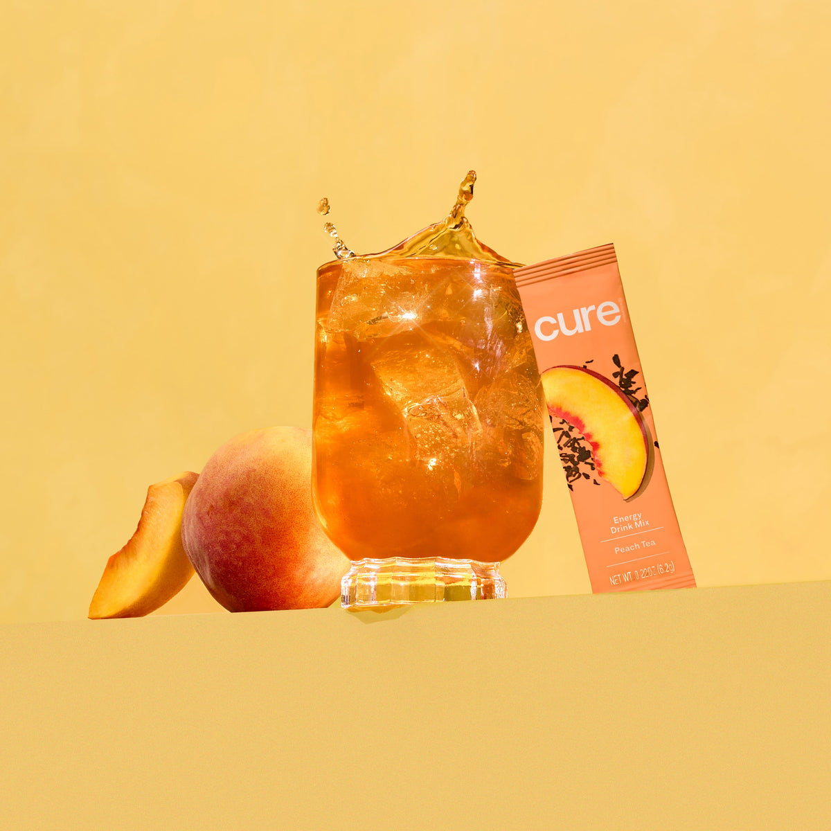 Glass of iced peach drink with a peach fruit, a peach slice, and a peach-flavored product box labeled 'cure'