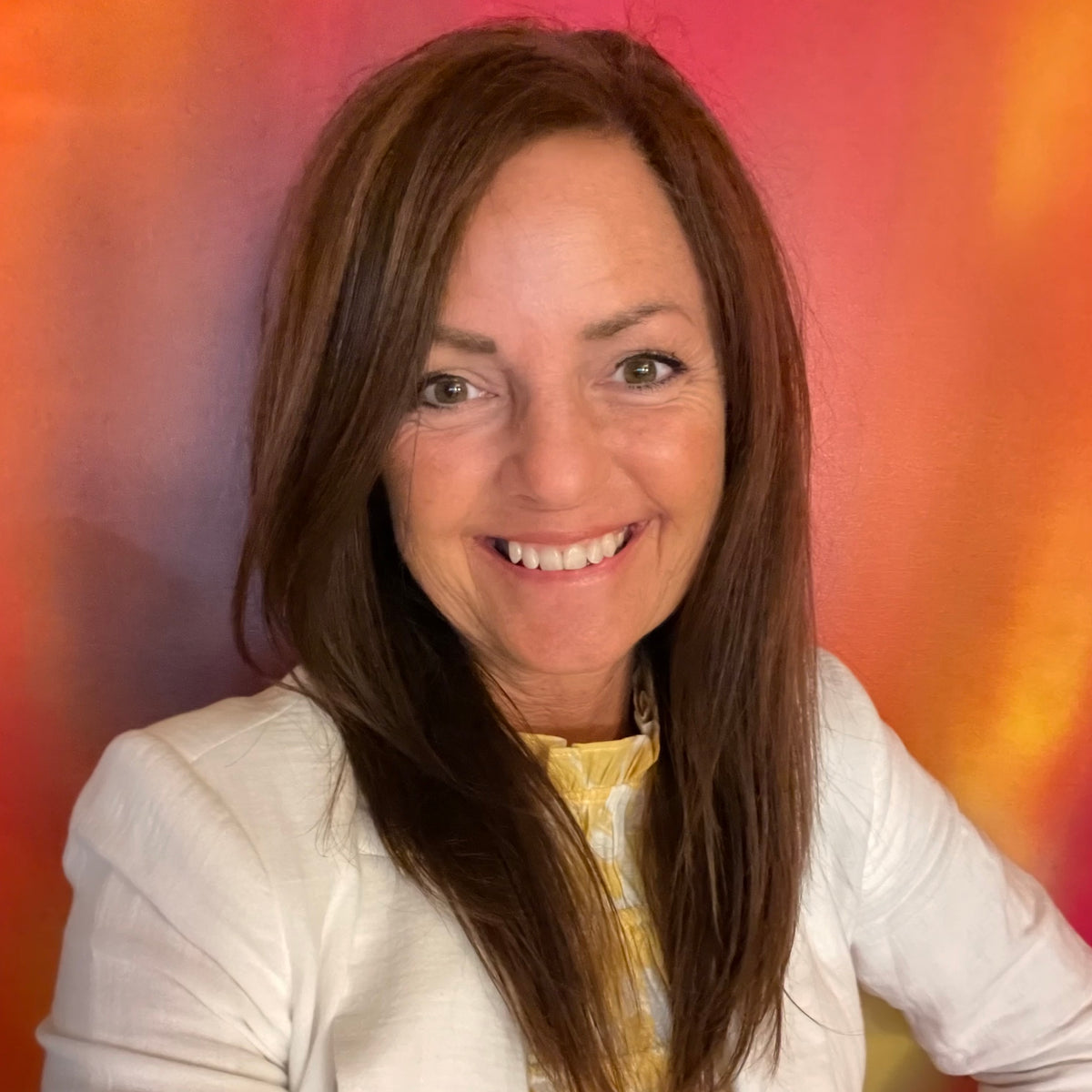 Smiling woman with long brown hair wearing a white jacket and yellow top against an orange-red background