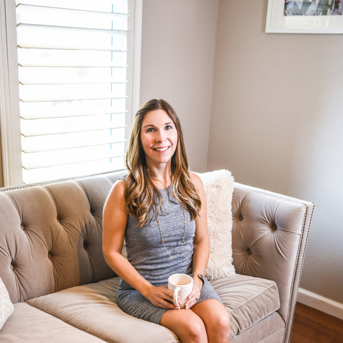 Woman with long hair in a gray dress sitting on a beige couch holding a white mug