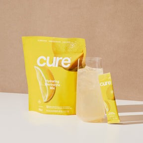 Yellow 'cure hydrating electrolyte mix' packet with lemon slice image, a glass of lemonade, and a single stick pack labeled 'cure'