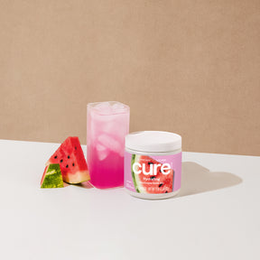 Jar labeled 'cure watermelon' next to a pink iced drink and two watermelon slices on a white surface.