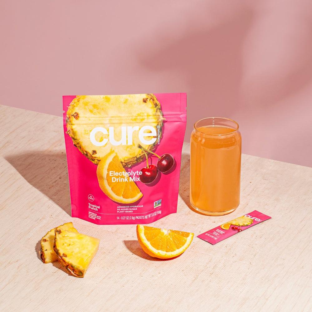 Cure brand Tropical Punch/Berry Pomegranate Bundle packaging with pineapple slice, cherries, a glass of orange drink, pineapple and orange fruit slices, and a single stick pack on a light surface with pink background