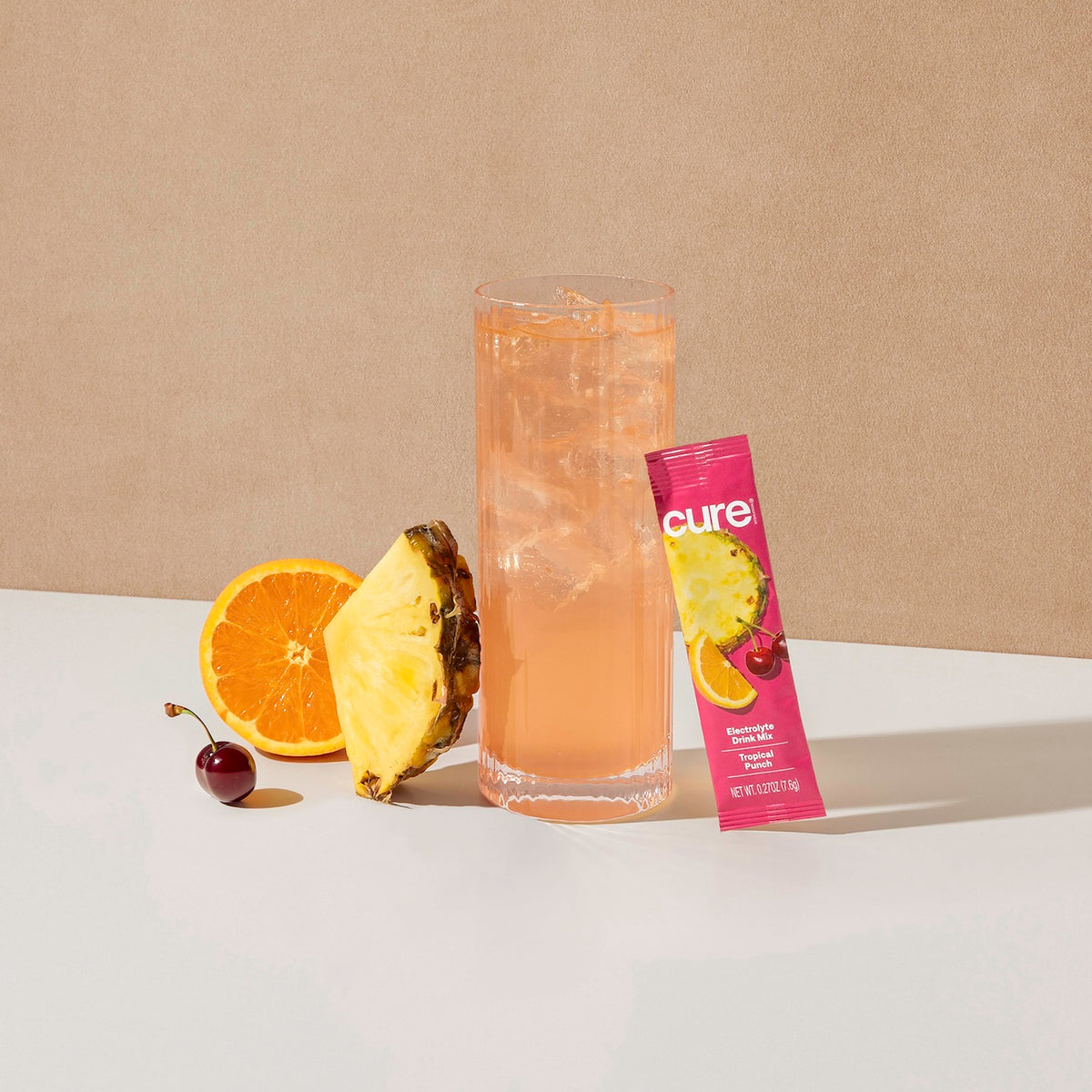 Glass of pink Tropical Punch drink with a Cure brand packet, orange slice, pineapple slice, and cherry on a white surface.