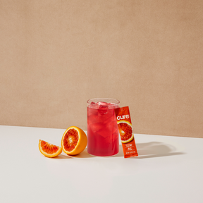 Glass of red tropical drink with blood orange slices and a Cure brand packet labeled 'cure'