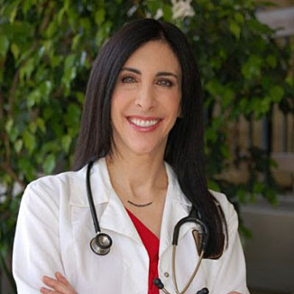 Smiling female doctor with long dark hair wearing a white coat and stethoscope, standing with arms crossed in front of green foliage.